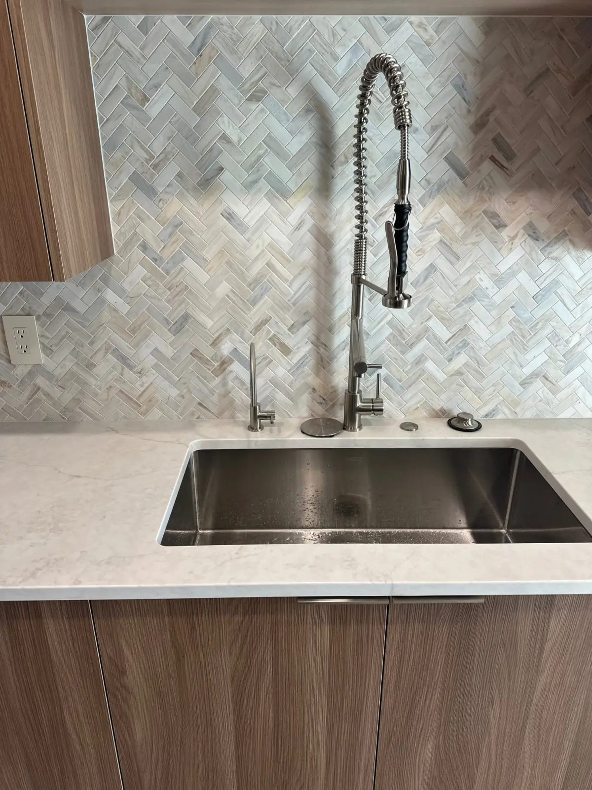 Stainless kitchen sink and faucet against a herringbone tile backsplash